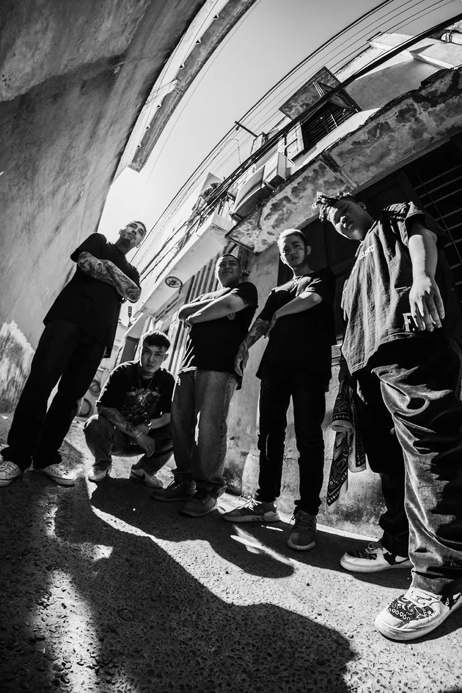 Five men in black tees in a sunlit alley, one holding a paisley bandana